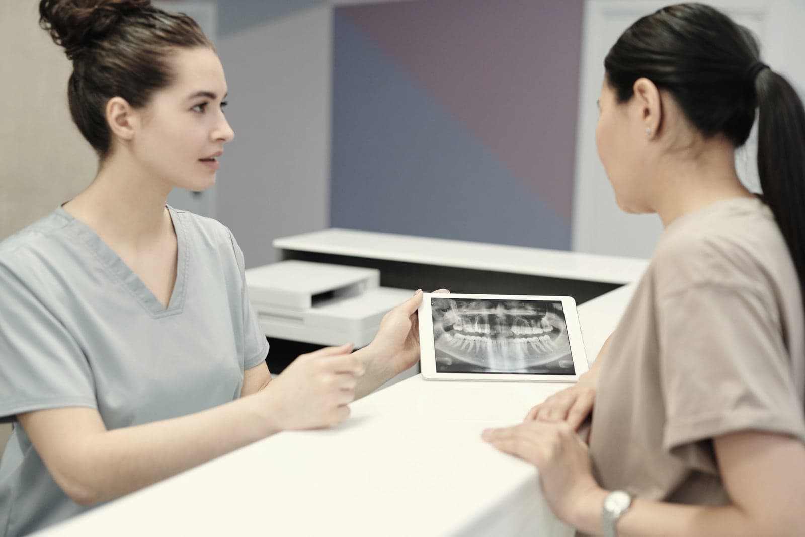 Across a desk, a dentist talks a patient through an X-ray on a tablet.