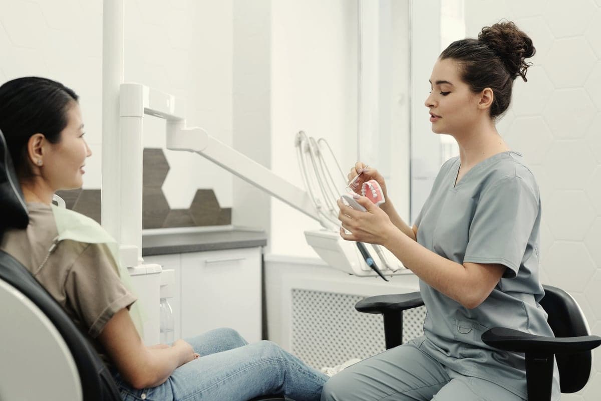 A dentist sits at eye level with a patient, explaining a treatment using a model of teeth.