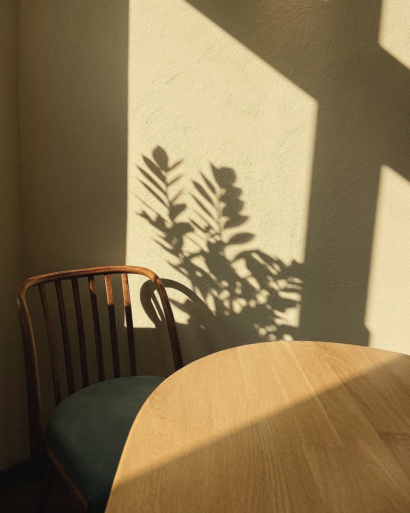 A wooden chair and table in a quiet, sunlit corner, with leaf shadows on the wall.