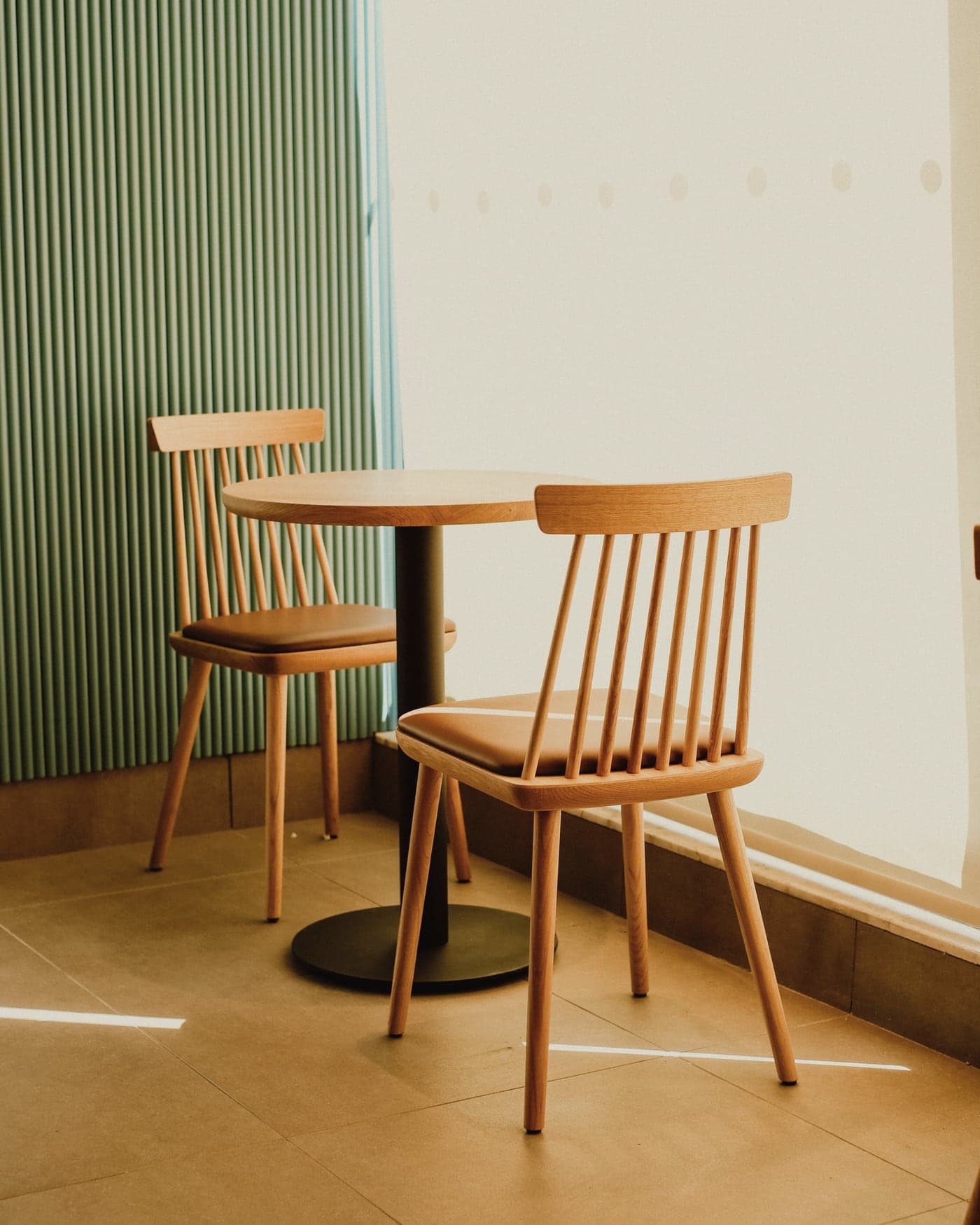 Two wooden chairs and a small table by a bright window, beside a sage-green slatted wall.